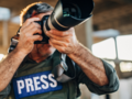 Ein Fotograf mit einer Weste und der Aufschrift "Press" schaut durch eine Spiegelreflexkamera