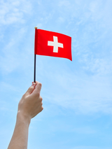Hand hält eine kleine Schweizer Flagge vor blauem Hintergrund