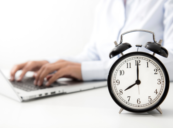 Ein Mensch tippt auf einem Laptop und eine Uhr steht im Vordergrund.