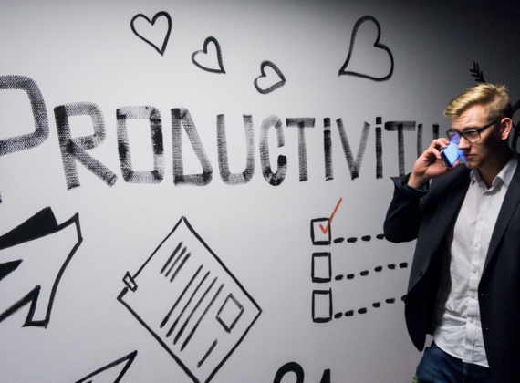 Ein junger Mann in Anzug telefoniert vor einer mit dem Wort Producvtivity beschrifteten Wand.