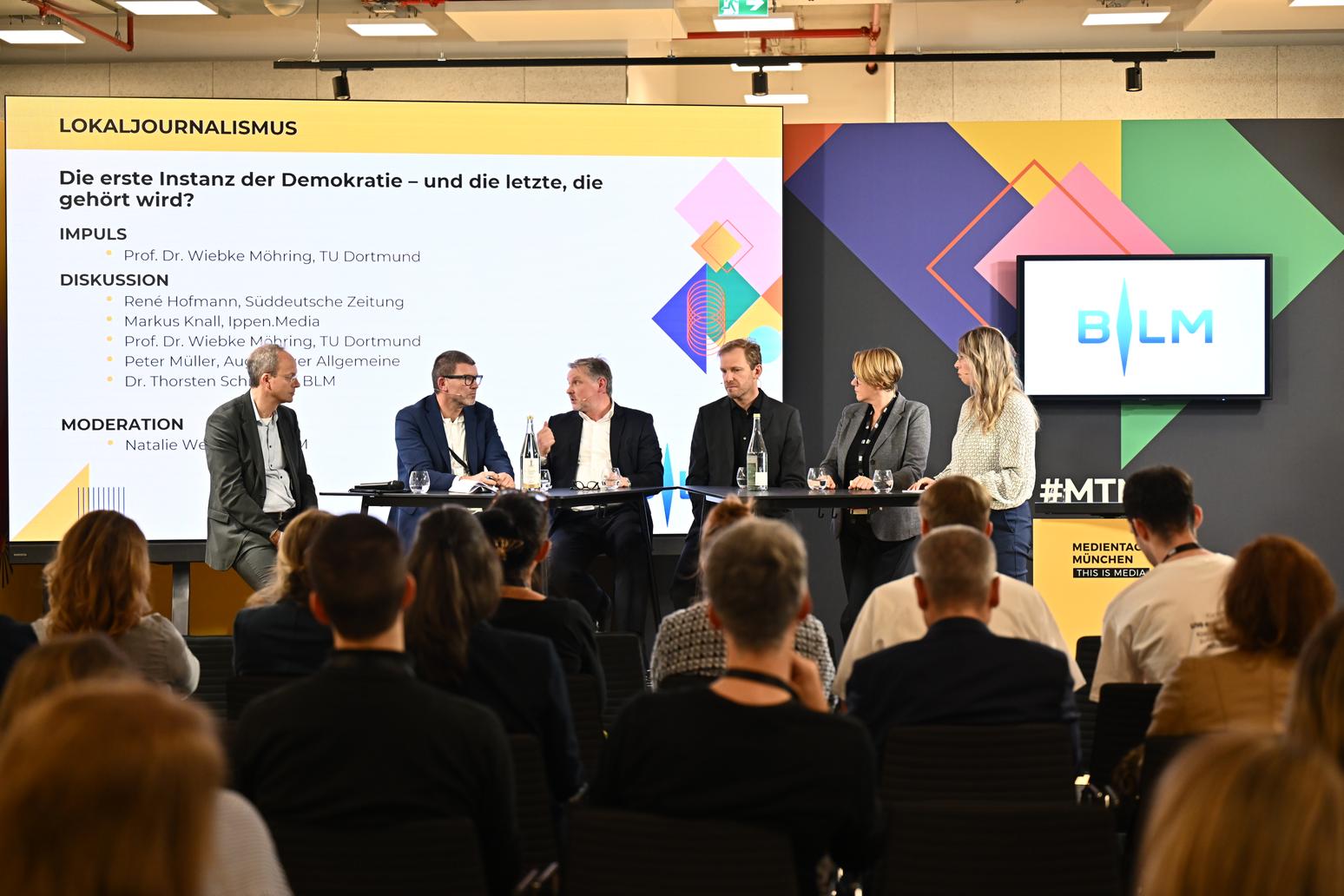 Podiumsdiskussion mit Experten aus Journalismus und Medien über die Bedeutung des Lokaljournalismus als demokratische Instanz. Im Hintergrund eine Präsentation mit dem Thema und bunte grafische Elemente. Publikum verfolgt aufmerksam die Diskussion.