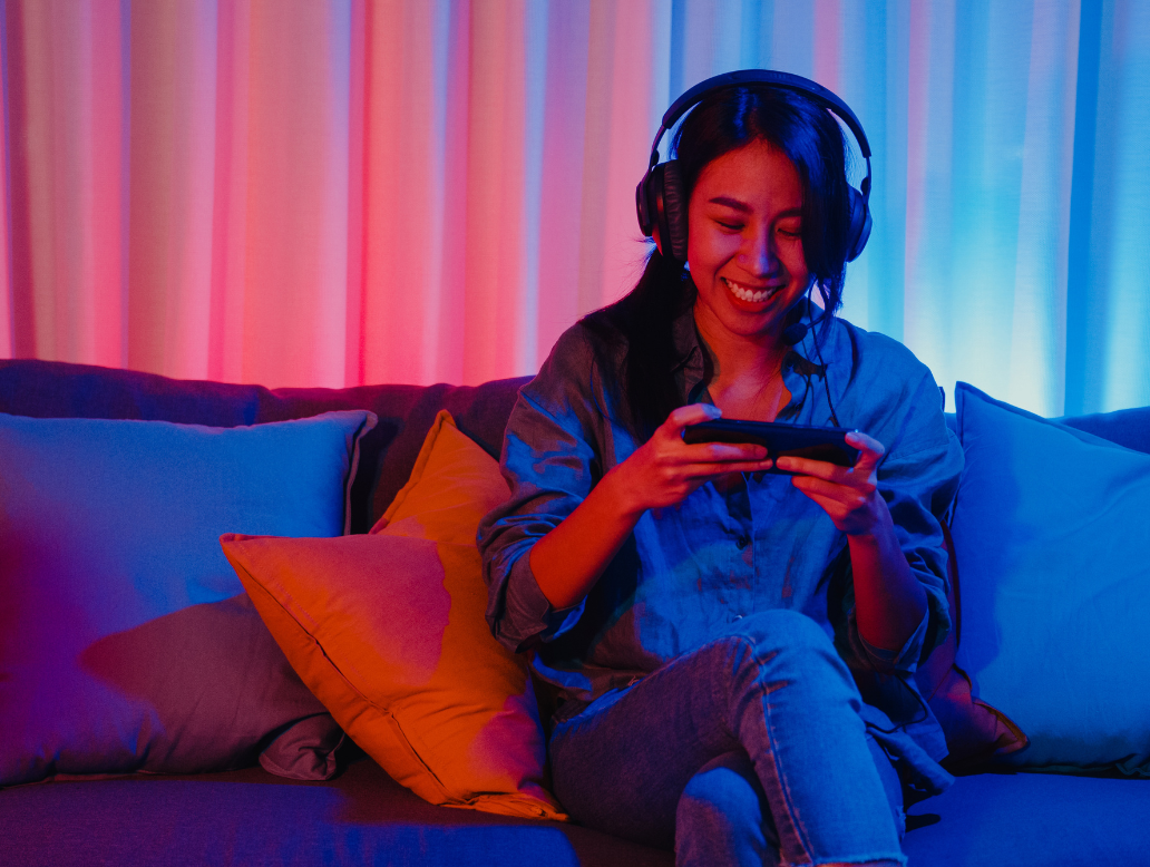 Happy asia Girl-Gamer tragen Headphone-Wettbewerb Video-Spiel online mit Smartphone aufgeregt Gespräche mit Freund sitzen auf der Couch in bunten Neonleuchten Wohnzimmer zu Hause, Home Quarantine Aktivität Konzept.